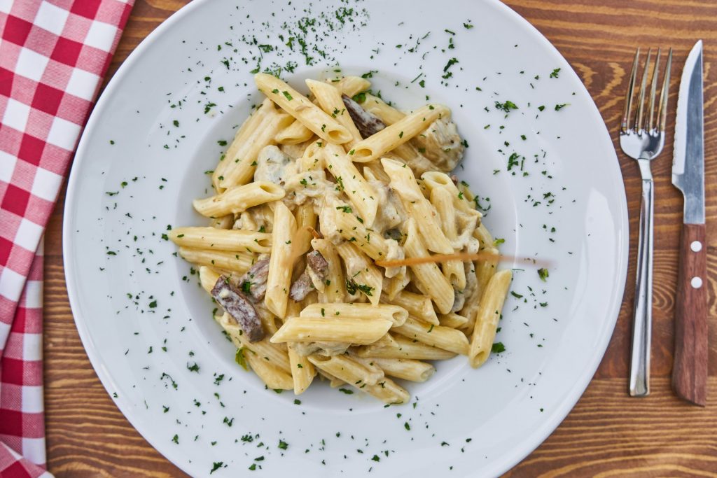 A top view of white plate of macaroni with seasoning on wooden table, fork and knife on the right side, and red and white chess colored kitchen cloth on the left side