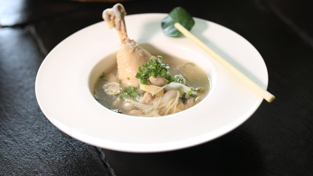 A white bowl of chicken soup with chicken leg, noodles in it, standing on the table