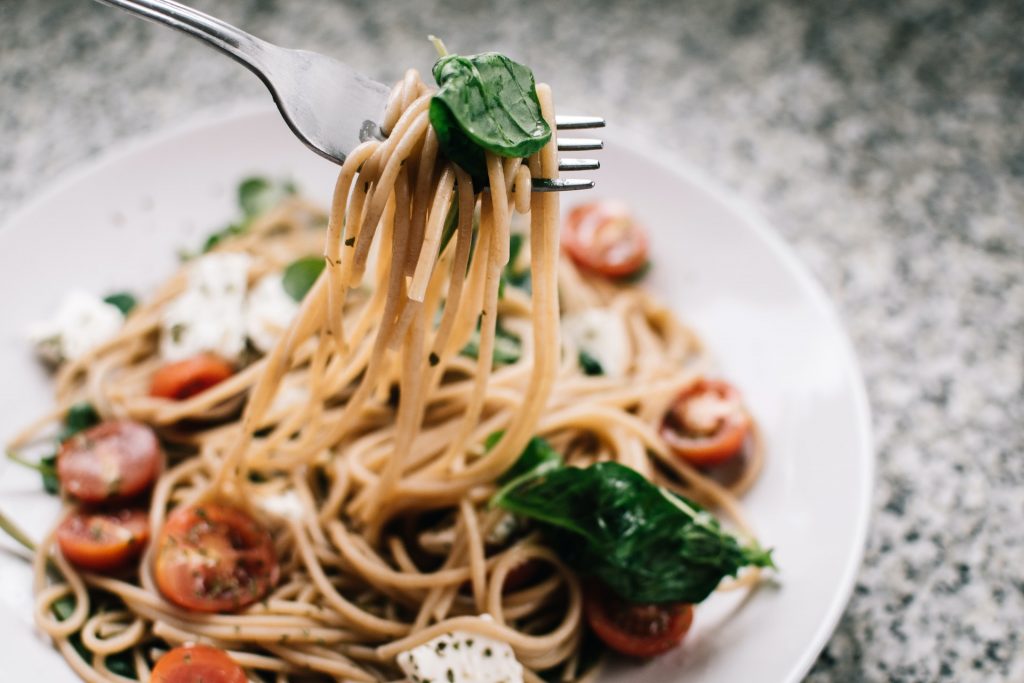 A fork holding a chunk of spaghetti above white plat with spaghetti on it, on the gray table top.