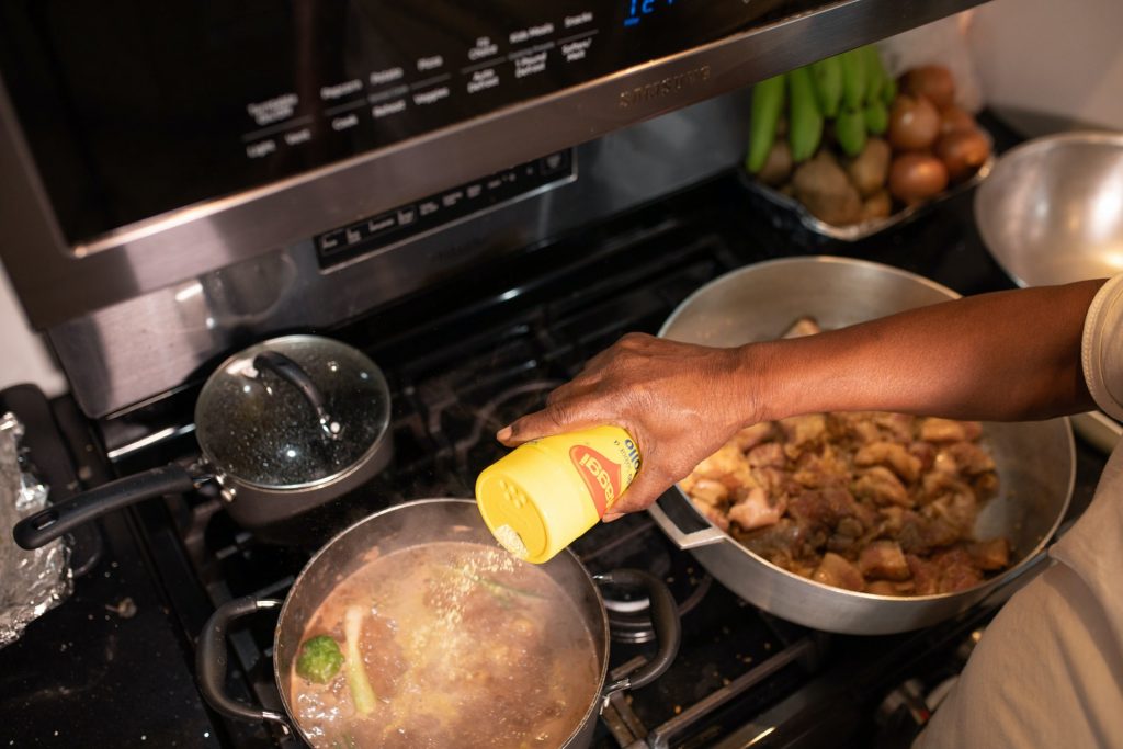 A hand of a cook pouring some powdered cheese into a pot with boiling chicken soup on a stove.