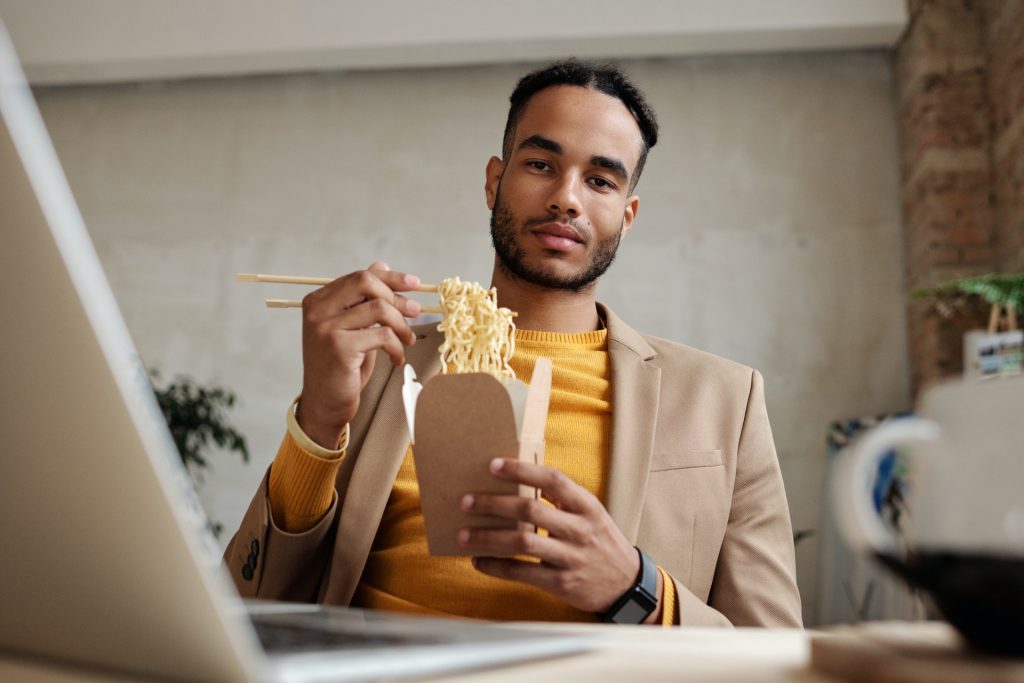 A Gentleman in a beige jacket and yellow sweater is eating a pack of ramen noodles by the table with laptop on it on beige wall background