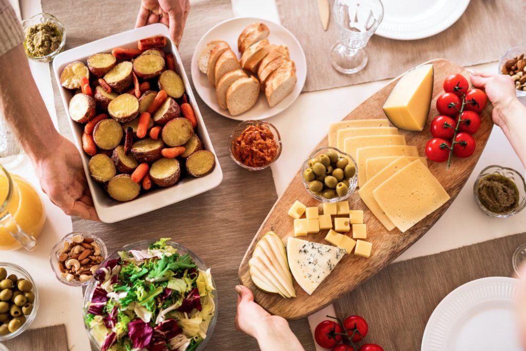 Someone's hands holding a tray with a variety of cheeses over the served table with bowls of potato, bowl of crackers, a salad, and so on. how to serve different types of cheeses