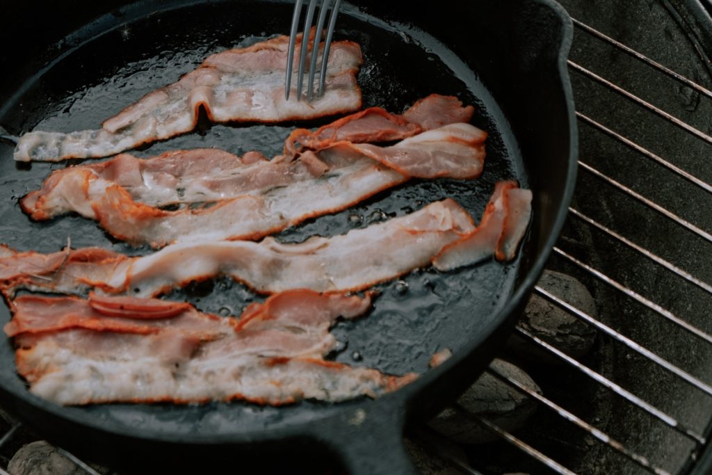 A batch of bacon grilling on a black metal pan on a grill grates