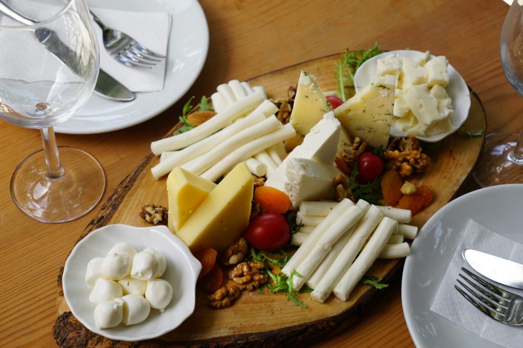 A cheese tray with several type sof cheeses and dips on it, standing on the brown tabletop along with two white ceramic plates with knifes and fork on each of them - How to Serve 18 Different Types of Cheeses