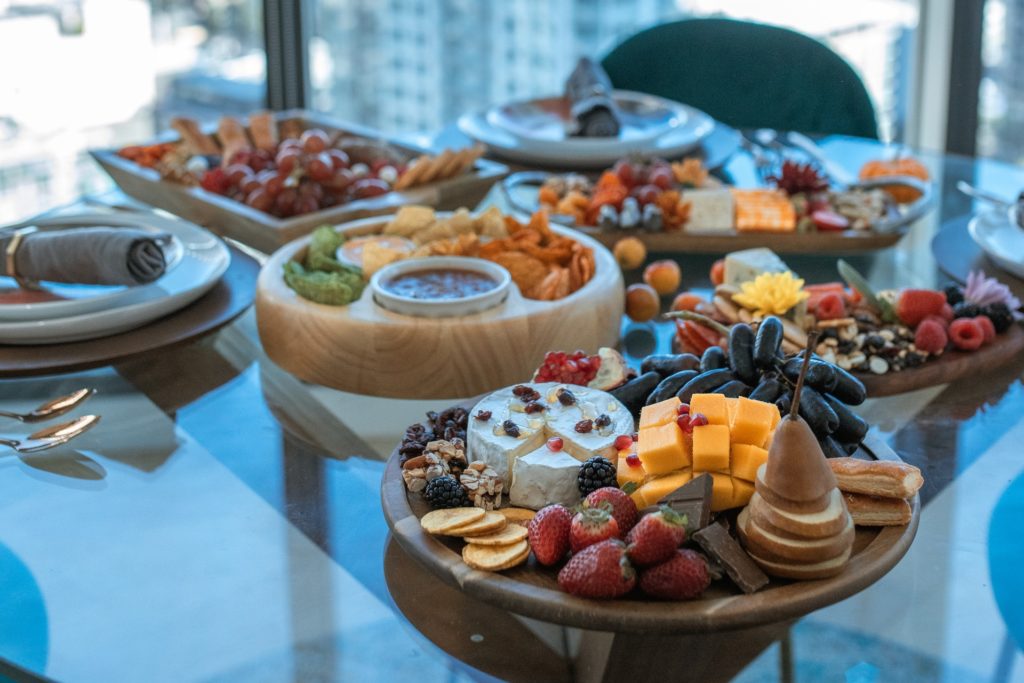 A cheese bowl with camembert cheese and some semi hard cheese pieces with strawberries, crackers and bowls many accompanying foods on glass standing table top. In the background nice city view from the high-rise building this is located at.