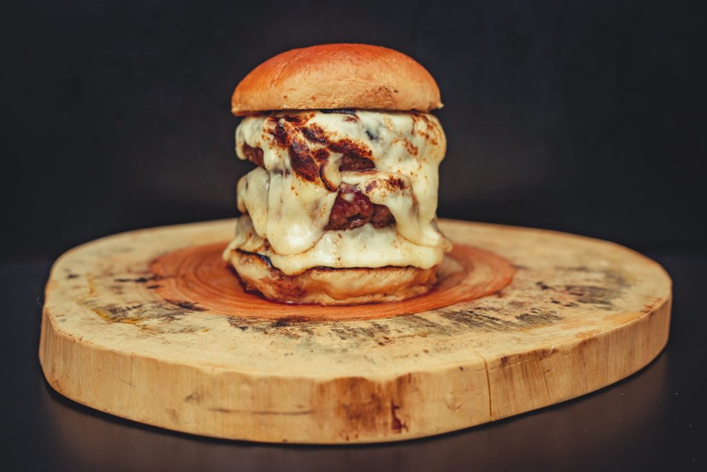 A burger with abundance of cheese sauce all over, on a wooden plate on black background.