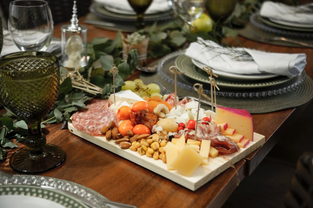 A cheese tray with pieces of hard and soft cheese, tomatoes, salami cuts, nuts served standing on a table top besides other clean dishes.